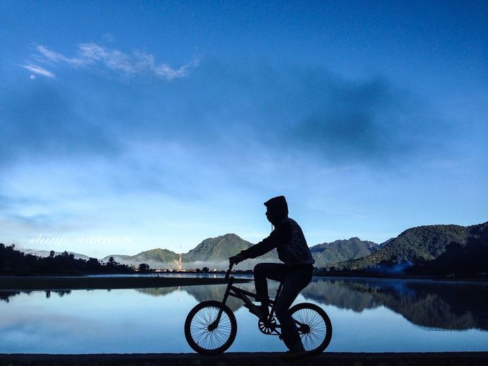 Silhouette man riding bicycle by lake against sky