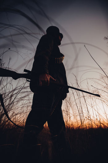 Low angle view of hunter with shotgun walking in field during sunrise