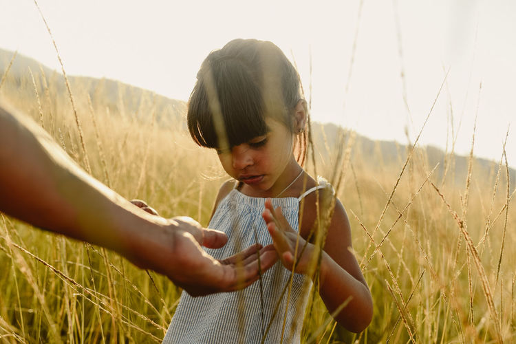 Unrecognizable crop adult parent extending hand to little girl against blurred of green meadow in daylight
