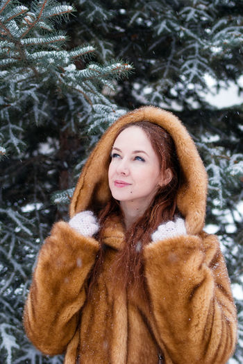 Portrait of young beautiful woman with red hair in faux fur coat on background of winter snowy park.