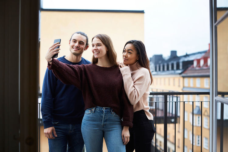 Smiling woman taking selfie with young friends while standing in balcony at rental apartment