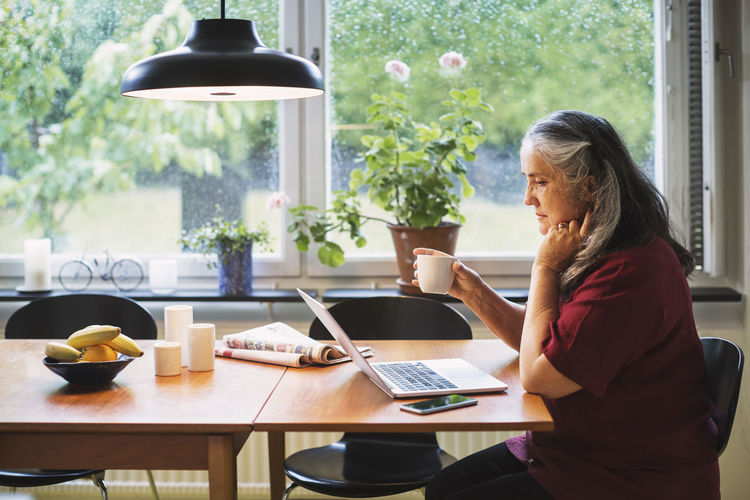 Side view of senior woman using laptop while having coffee at table