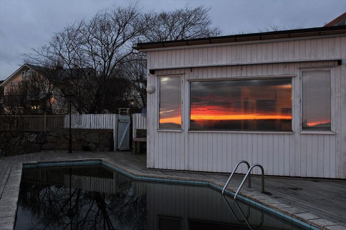 Swimming pool by house with reflection of orange sky on glass window