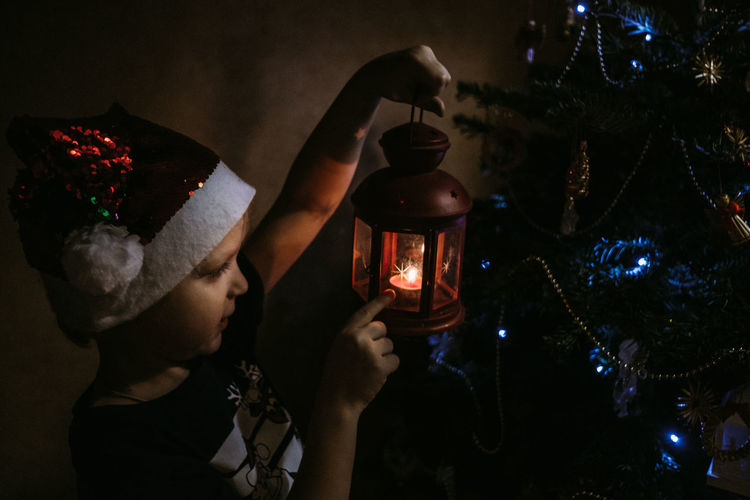 A child in santa's hat is near illuminated christmas tree at night