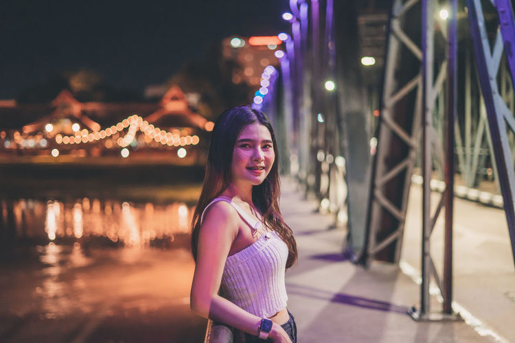 Portrait of smiling young woman standing on bridge over river at night