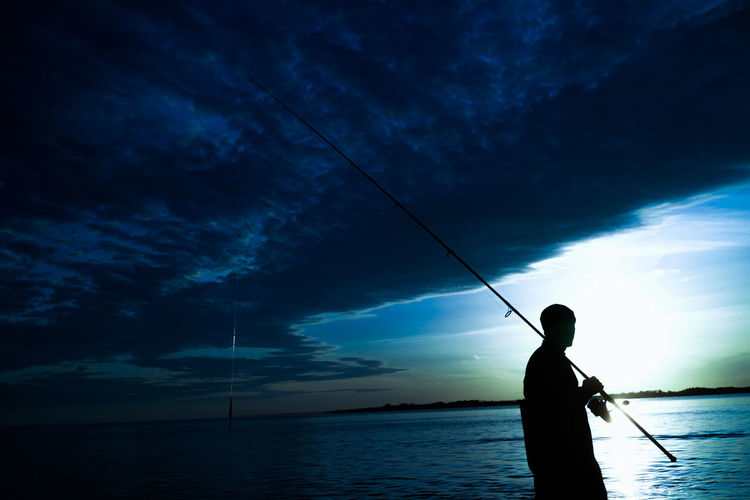Silhouette man fishing in sea against cloudy sky