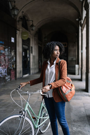 Black woman in smart casual style walking along street with bike and looking away