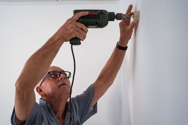 Concentrated old male in eyewear with electric screwdriver screwing plastic piece to wall in house
