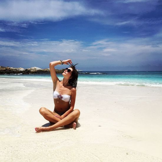 Young woman in bikini sitting on beach against sky
