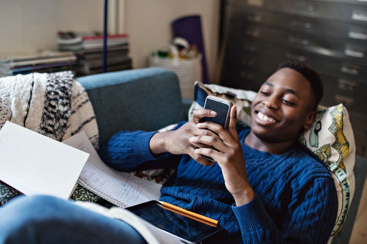 Smiling teenage boy using social media while lying on sofa during homework in living room