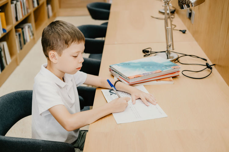 The boy sits in the library and writes in a notebook at the table. preparing for homework. 