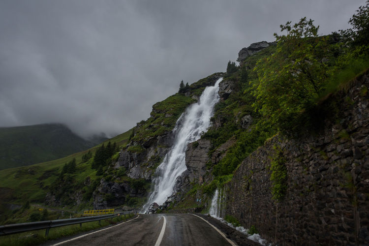 Road leading towards waterfall against cloudy sky