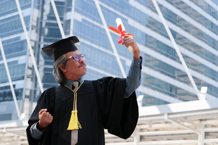 Businessman in graduation gown clenching fists against office building