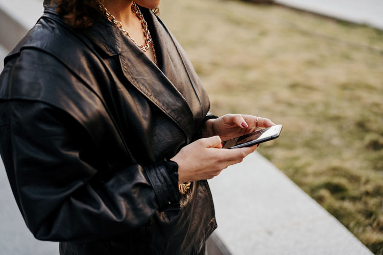 Young businesswoman in retro black leather jacket and skirt browsing smartphone while standing in arched passage in park before work