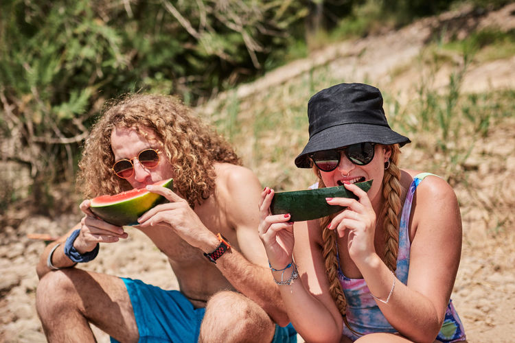 Company of friends sitting on tree log in forest and eating delicious watermelon while enjoying summer holiday together