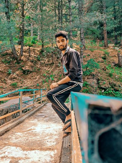 Indian young man sitting on iron bridge background pine tree wearing black t-shirt beauty in nature.