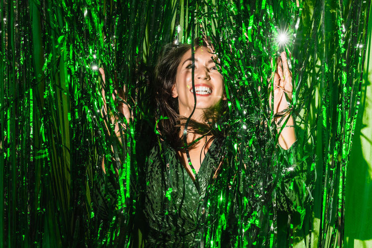 Delighted female wearing festive dress playing with green shiny tinsel stripes at party while looking at camera