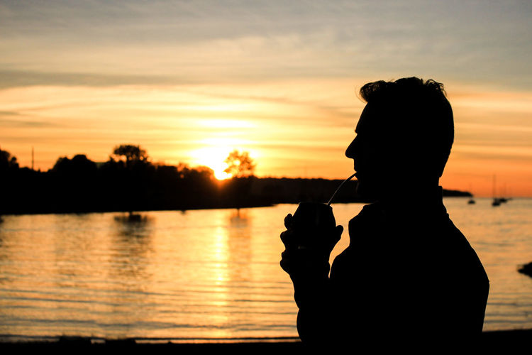 Side view of silhouette man drinking by lake during sunset