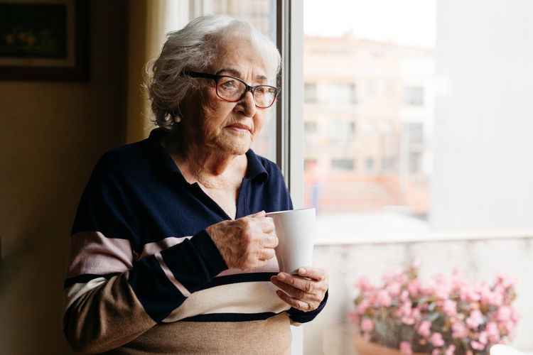 Side view of elderly female standing near window with cup of coffee and enjoying morning at home