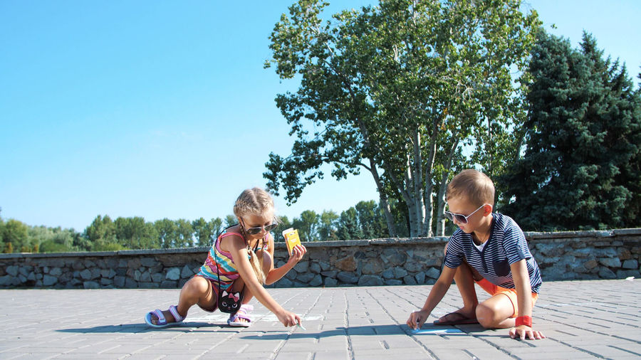 Children, a boy and a girl in sunglasses, paint with colored crayons on the asphalt, street tiles