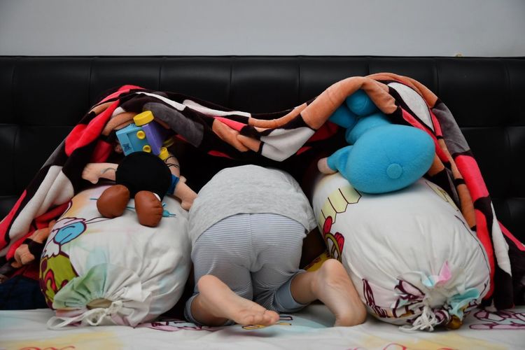 Child hiding with toys below blanket on bed at home