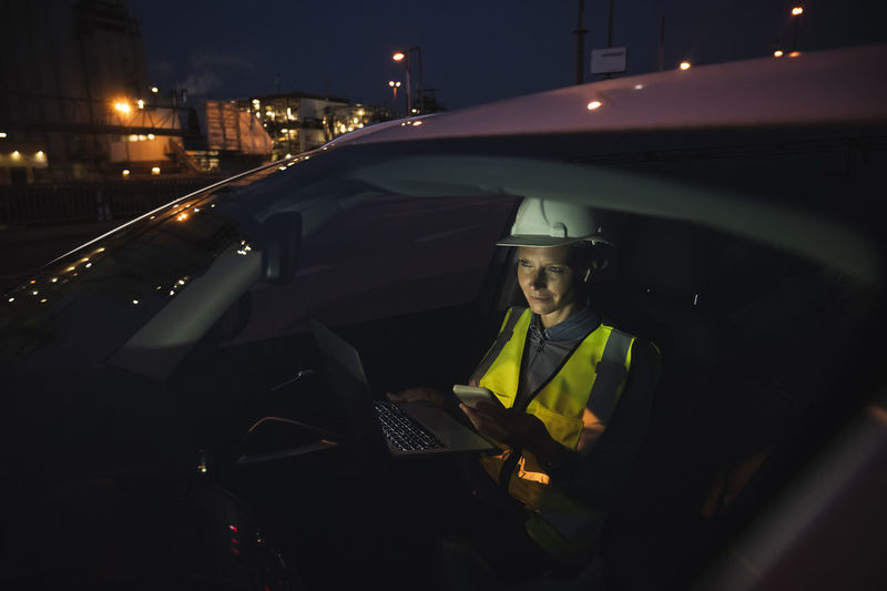 Female professional using technologies while sitting in car seen through windshield at night