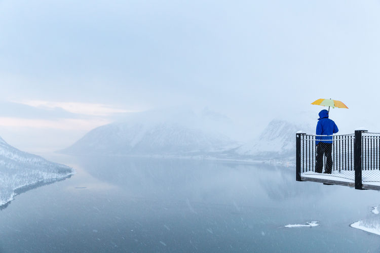 Rear view of man standing with umbrella at observation point over lake during snowfall