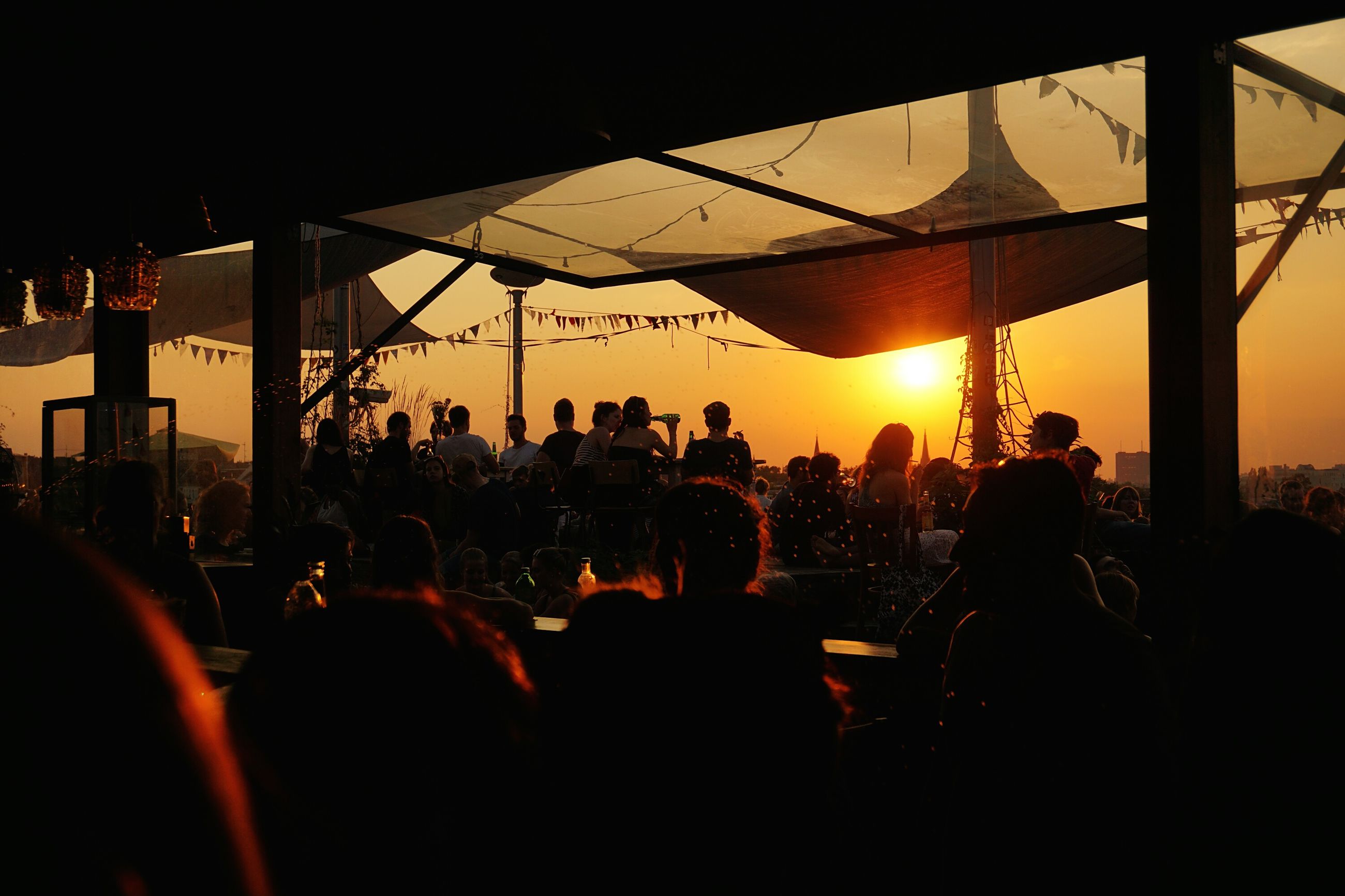 sunset, large group of people, silhouette, men, person, lifestyles, sun, leisure activity, sky, orange color, built structure, architecture, city, sunlight, enjoyment, mixed age range, sea, medium group of people, city life