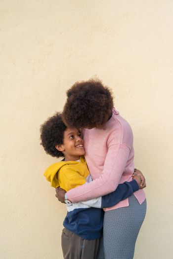 Side view of tender african american woman and boy embracing on street and looking at each other while standing in city against beige wall