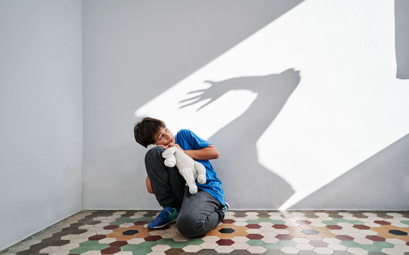 Scared little boy with toy in hands sitting near wall with shadow of angry violent parent with raised arm