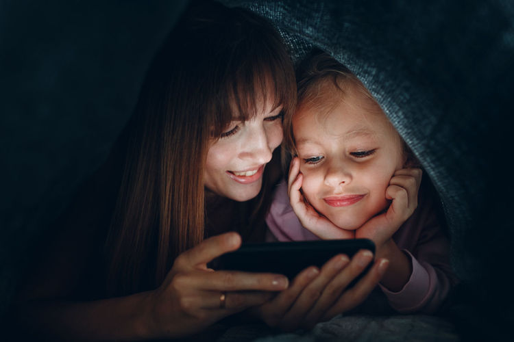 Smiling mother and daughter looking at mobile phone under blanket