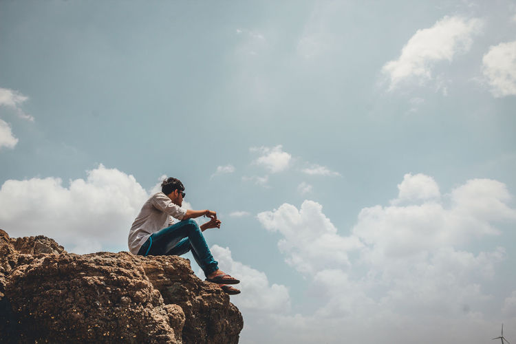 Low angle view of young man sitting on cliff against sky