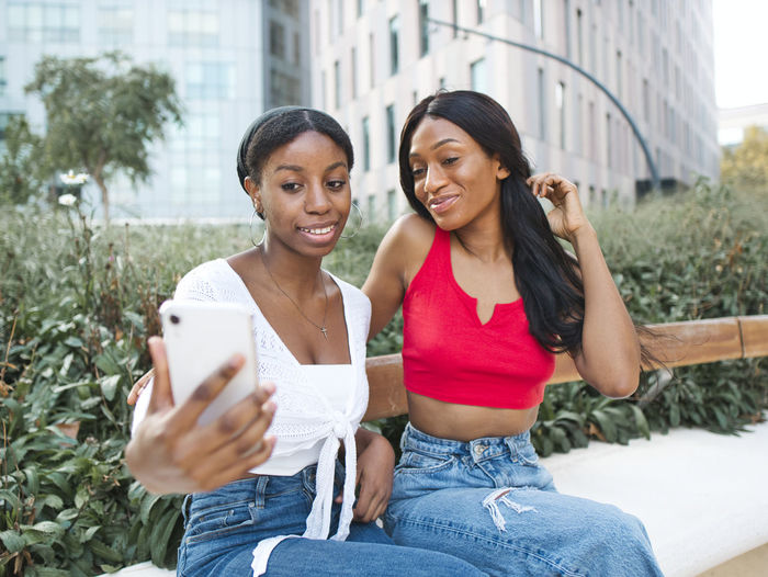 Delighted african american female friends taking self portrait on cellphone while sitting on bench near green plants on street with modern buildings