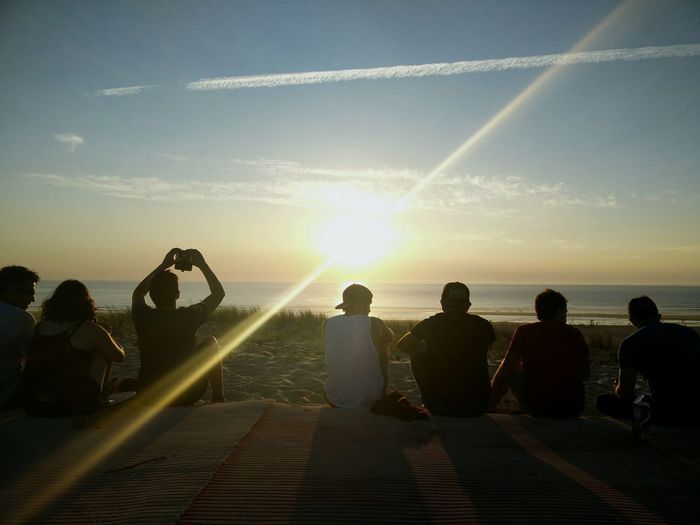 Rear view of friends sitting at beach against sky during sunrise