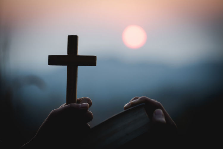 Low angle view of hand holding cross against sky during sunset