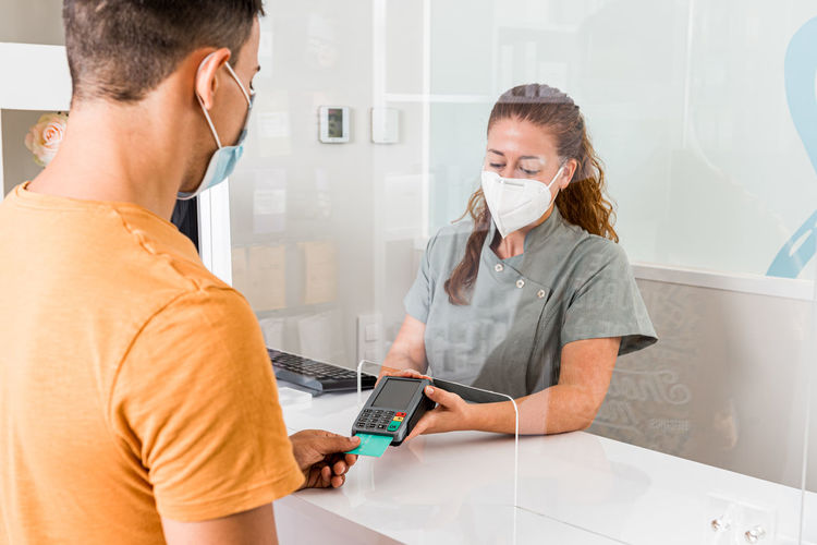 Young female specialist in protective mask holding card reader while male patient paying for treatment with credit card in modern clinic