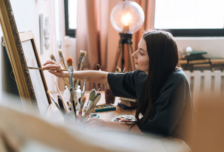 Young woman artist teenager girl student with dark long hair in casual draws picture at art studio
