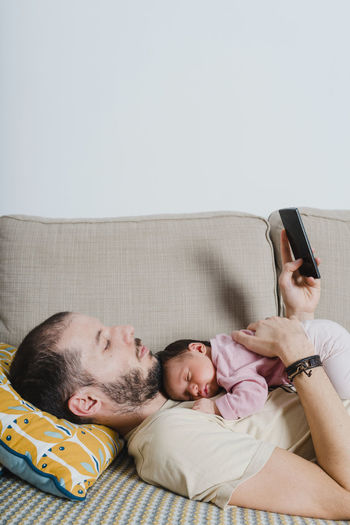 Father lying on the couch with newborn baby girl using smartphone