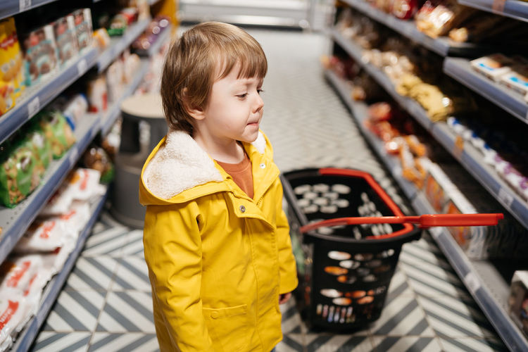 Child in the market with a grocery cart, wearing a yellow jacket and jeans
