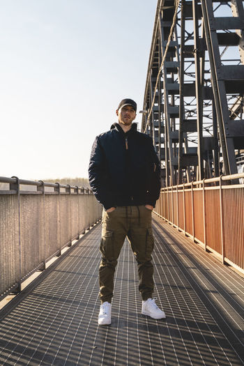 Portrait of young man standing on footbridge against clear sky