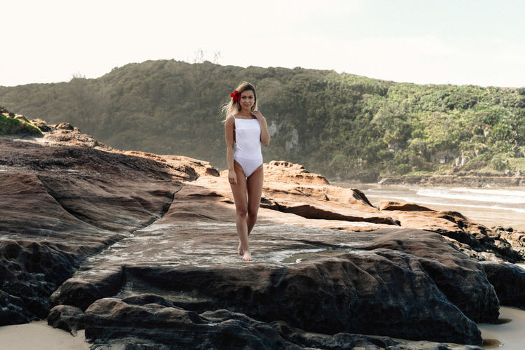 Seductive young woman wearing white swimsuit while standing on rock at beach