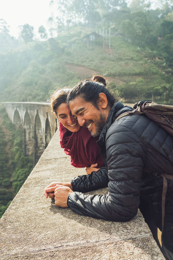 Side view of cheerful ethnic man and woman in activewear with backpacks talking and laughing while standing on aged stone nine arch bridge among green hills in sri lanka