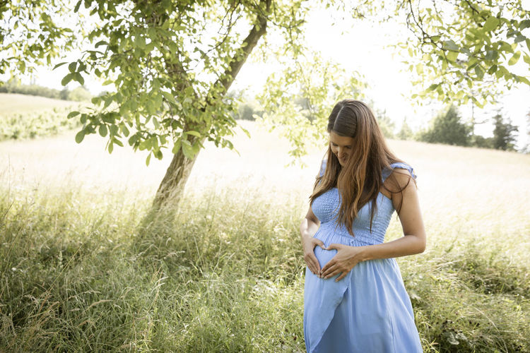 Pregnant woman making heart shape on stomach standing in meadow