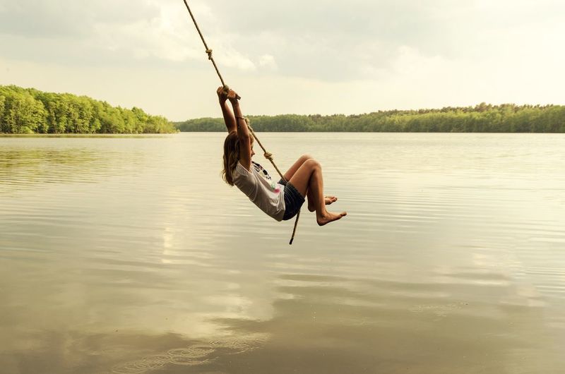 Girl swinging on rope over lake against sky