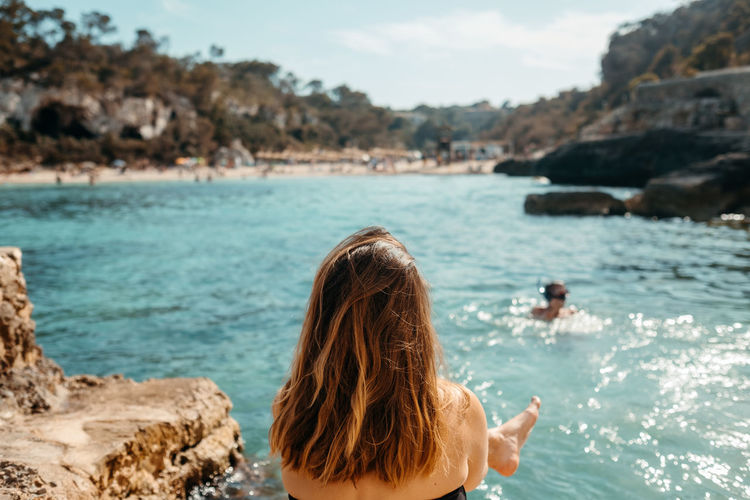 Back view of unrecognizable young female traveler with long wavy hair in swimwear relaxing on rocky seashore and admiring nature during summer holidays in mallorca