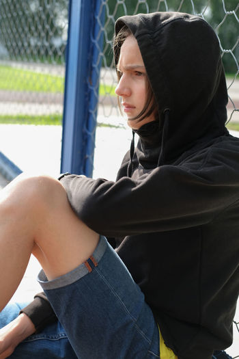 Handsome teenager boy sitting near net fence. teenager wearing black hoodie with hood. . sad guy. 