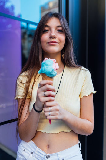 Crop cheerful young female in pendant and earrings with delicious gelato in waffle cone looking at camera on street