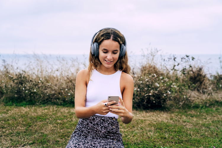 Enthusiastic woman in headphones placing on brick ledge in grass and listening to music from mobile phone