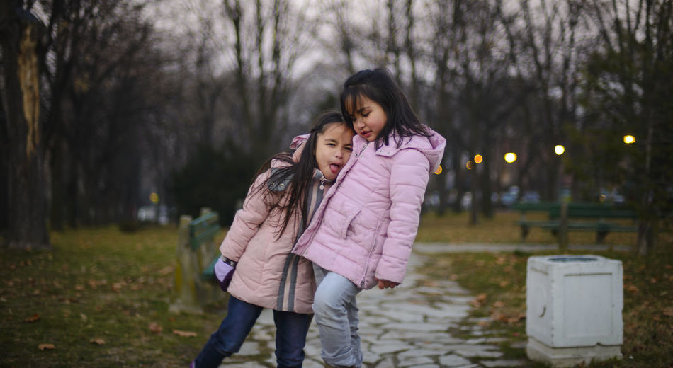 Playful sisters wearing pink jackets standing on pathway at park