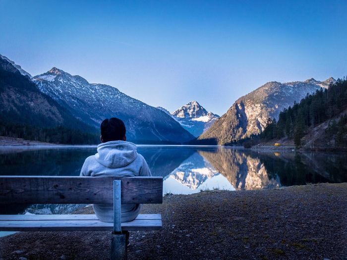 Rear view of man sitting at lakeshore against mountains during winter